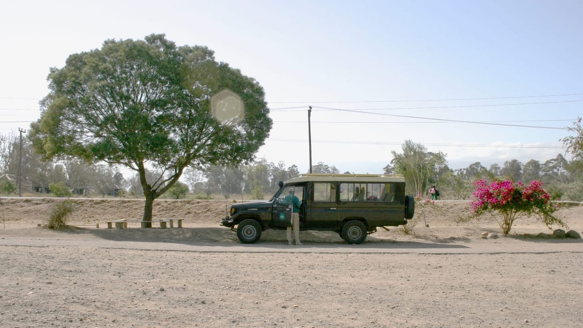 a dusty road on which is Parker a green jeep with a ranger tending through the passenger door. Trees lank the vehicle.