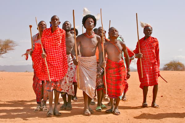 A tribe of topless tribal males approach dressed in red with eyes closed in ceremony.