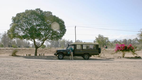 a dusty road on which is Parker a green jeep with a ranger tending through the passenger door. Trees lank the vehicle.
